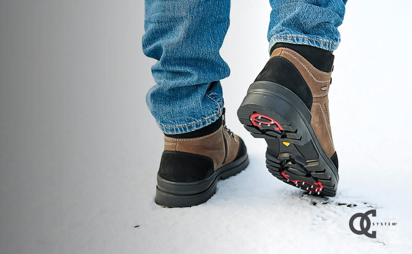 Person i jeans bär bruna och svarta vandringskängor med robust sula och vändbara dubbar, stående på snö.