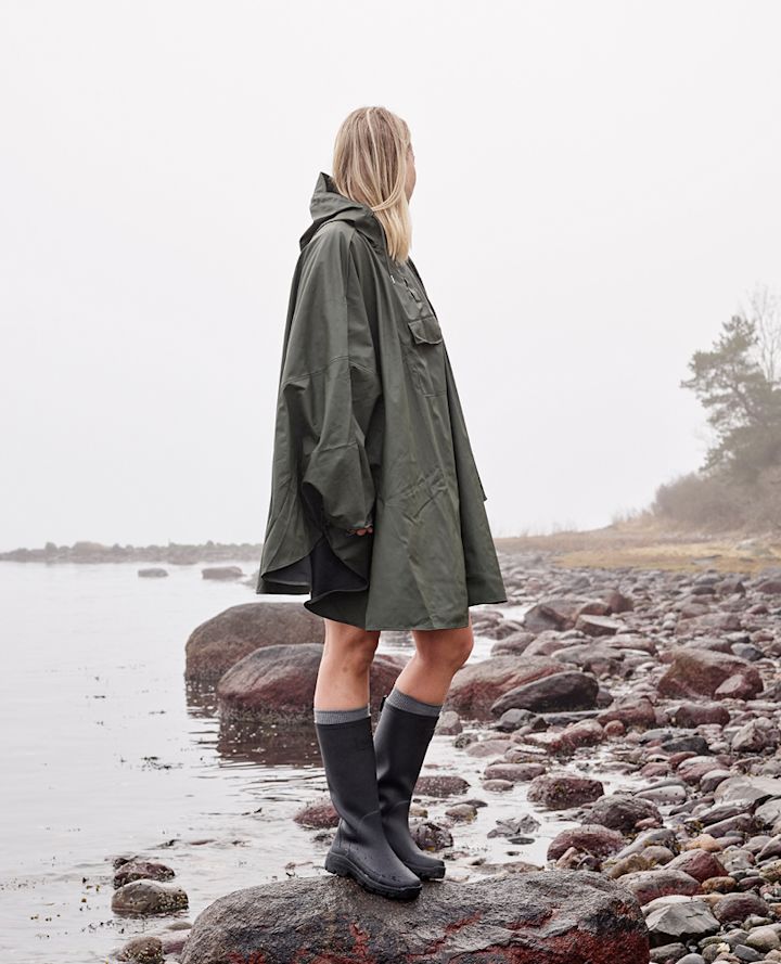 Person står på en stein ved vannkanten iført grønn regnponcho og svarte støvler, med ryggen til kameraet på en steinete strand.