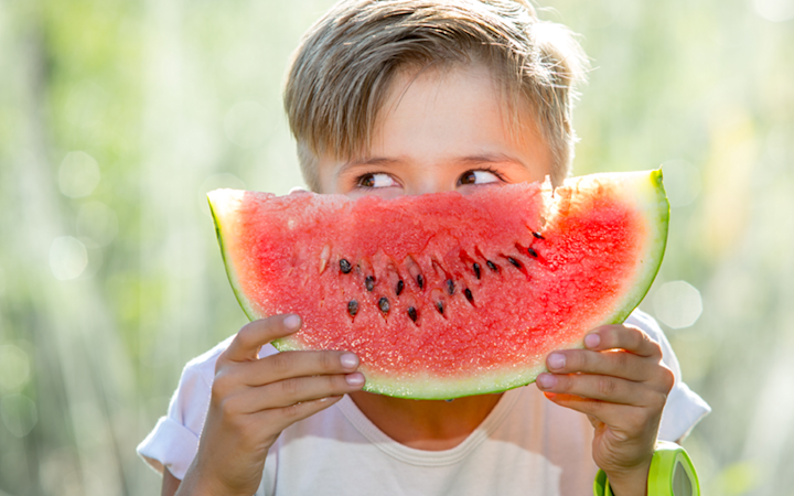 En pojke håller upp en stor skiva vattenmelon framför ansiktet och tittar åt sidan i en ljus utomhusmiljö.
