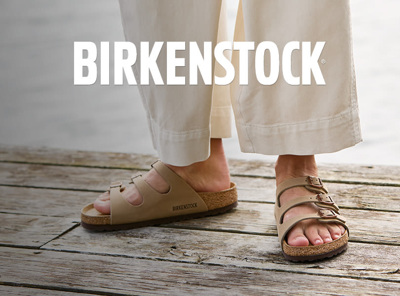 Beige Birkenstock sandaler med korksåle på tregulv, person med lyse bukser, merkelogo vises tydelig over bildet.