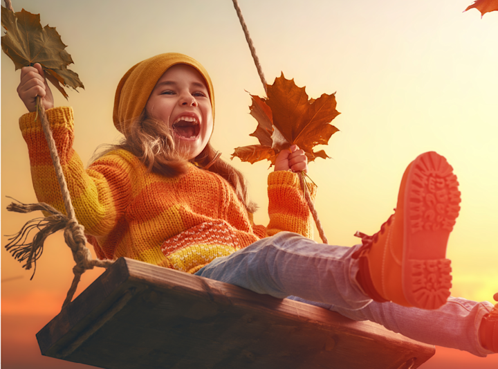 Liten jente i oransje og gul genser og lue svinger på en huske og holder høstløv, med solnedgang i bakgrunnen.