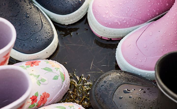 Flera par färgglada stövlar, inklusive rosa, svarta och blommiga, står i en vattenpöl med vattendroppar på sig.