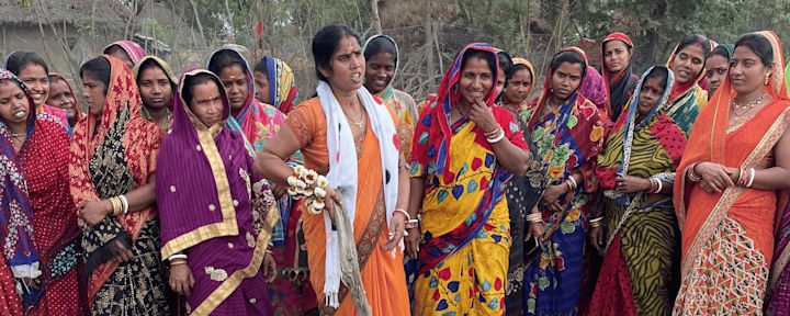 En grupp kvinnor i färgglada traditionella saris står tillsammans utomhus, några ler medan andra ser allvarliga ut, omgivna av träd och naturlig bakgrund.