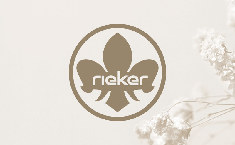Logo: Rieker tekst i en sirkel med en liljeformet figur i bakgrunnen, omgitt av tørkede blomster på høyre side.