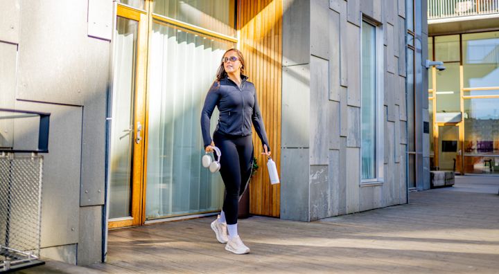 Person i svart träningsdräkt med vita löparskor i modern design går utanför en byggnad med trädetaljer, bärande hörlurar och vattenflaska.