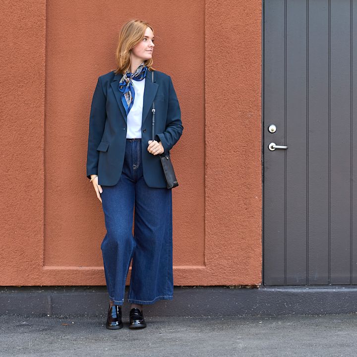 Person i mörkblå kavaj och vida jeans mot tegelvägg, bär blanka svarta loafers i klassisk stil.