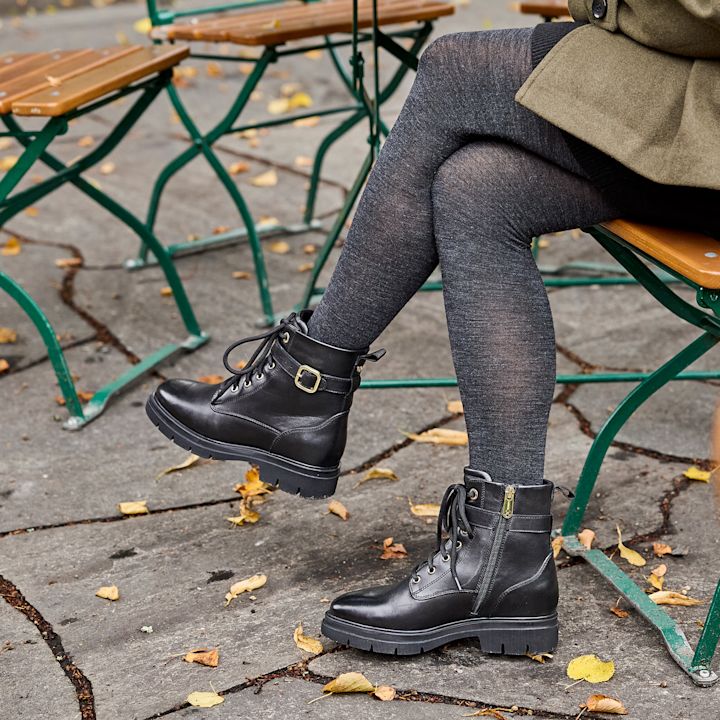 Svarta skinnst övlar med dragkedja och spänne på sidan, bära av en person med grå strumpbyxor som sitter på en bänk utomhus med höstlöv på marken.