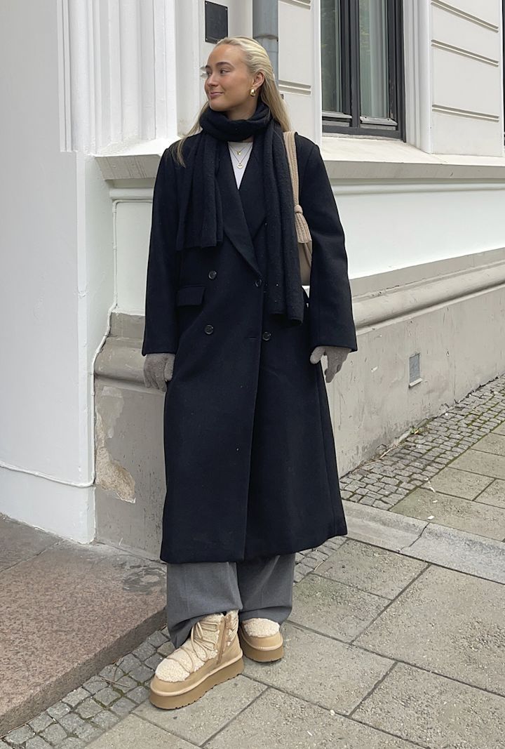 Kvinna bär en lång svart kappa, svart halsduk, gråa handskar, grå byxor och beigea vinterstövlar och står på trottoaren vid en vit byggnad med fönster.