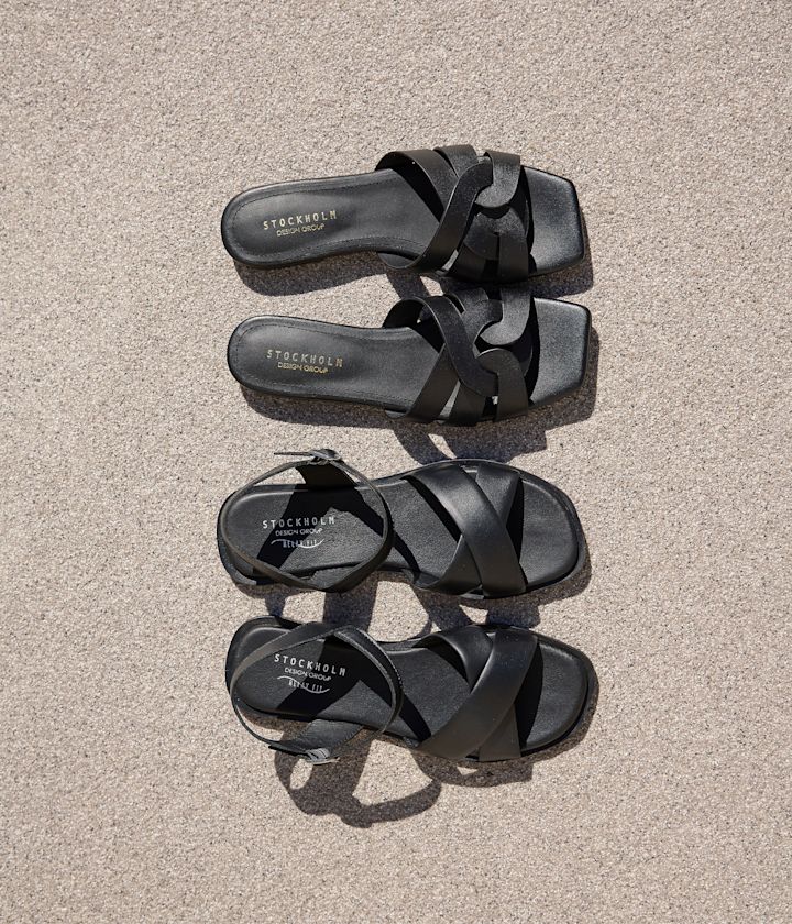 Två par svarta sandaler från Stockholm Design Group, en slip-in och en med ankelrem, fotograferade uppifrån på sand.