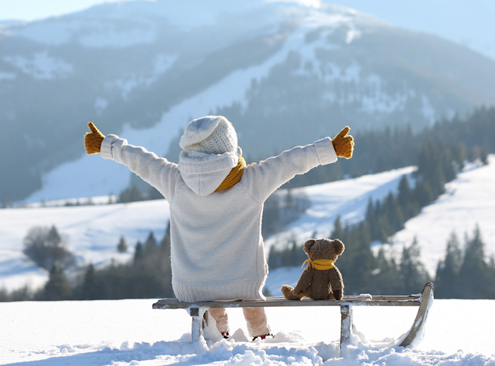 Barn i vit stickad tröja, mössa och gula vantar sitter på en kälke i snön med armarna utsträckta, bredvid en nallebjörn med gul halsduk, med snötäckta berg i bakgrunden.