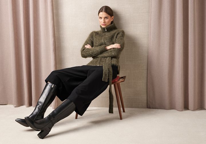 Modell i grønn genser og svarte bukser sitter på krakk, iført elegante svarte skinnstøvletter med hæl og glidelås.