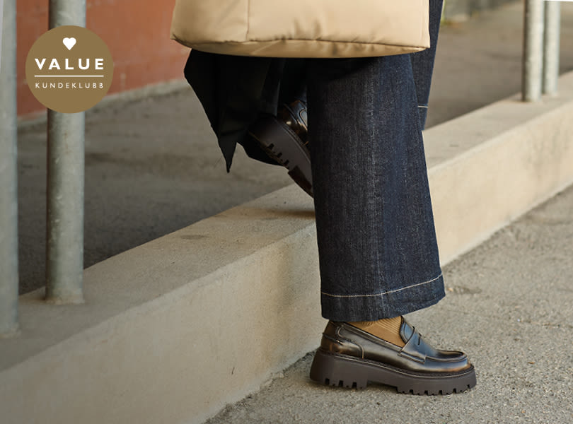 Svarte, klassiske loafers fra Unified med plattformsåle under mørke jeans. VALUE kundeklubb-logo vises i hjørnet.