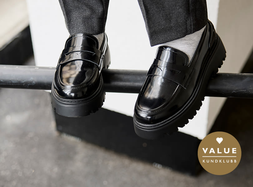 Svarta blanka loafers från Unified med kraftig sula, burna med gråa byxor och vita strumpor. Value kundklubb-logotyp i hörnet.