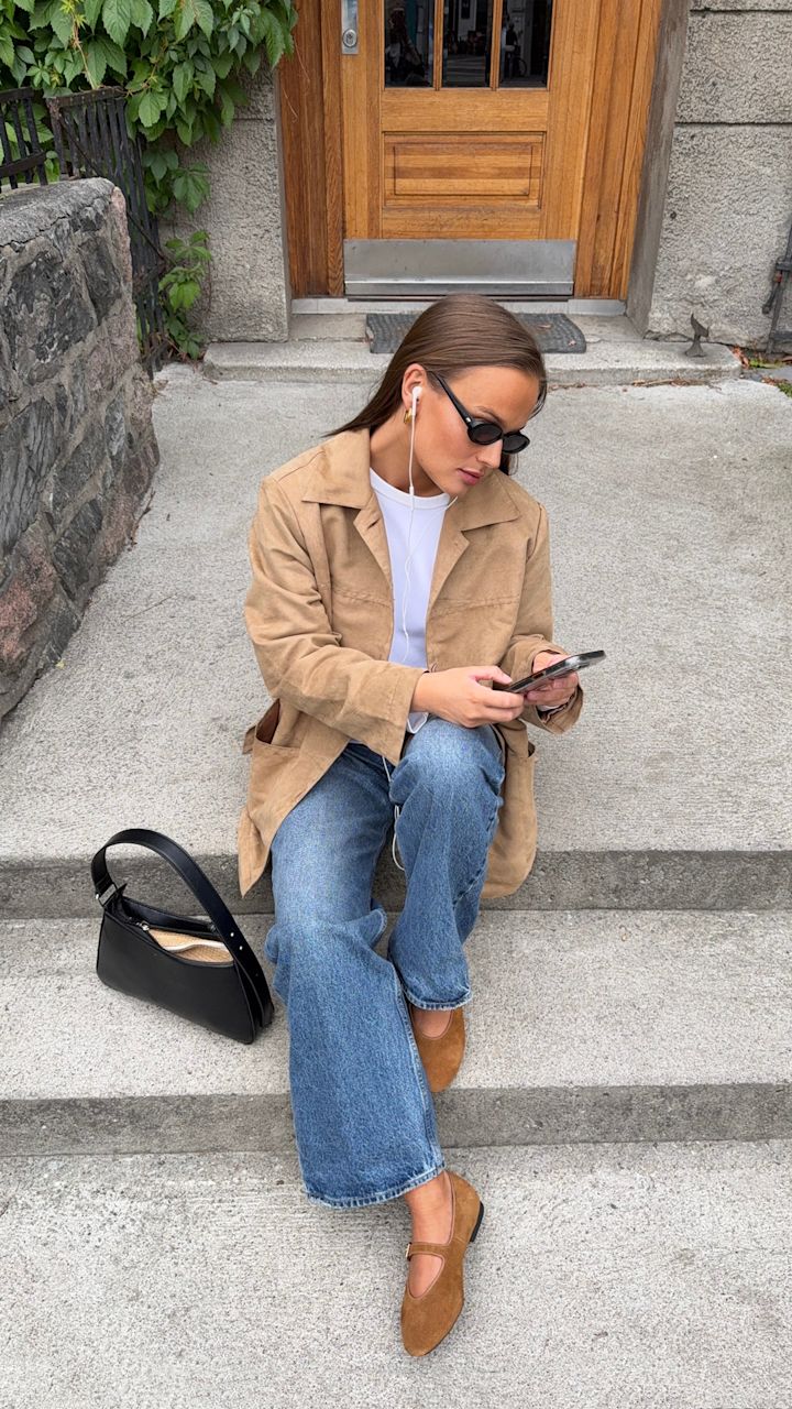 Kvinna med solglasögon och beige jacka sitter på en betongtrappa utanför en byggnad, använder öronproppar och tittar på mobilen med en svart väska bredvid sig.