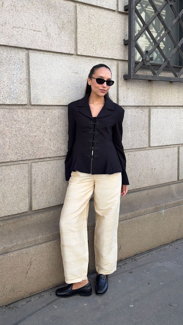 Ingrid Edvinsen i svart blazer og beige bukser poserer mot steinvegg, iført klassiske svarte loafers i skinn med lav hæl.