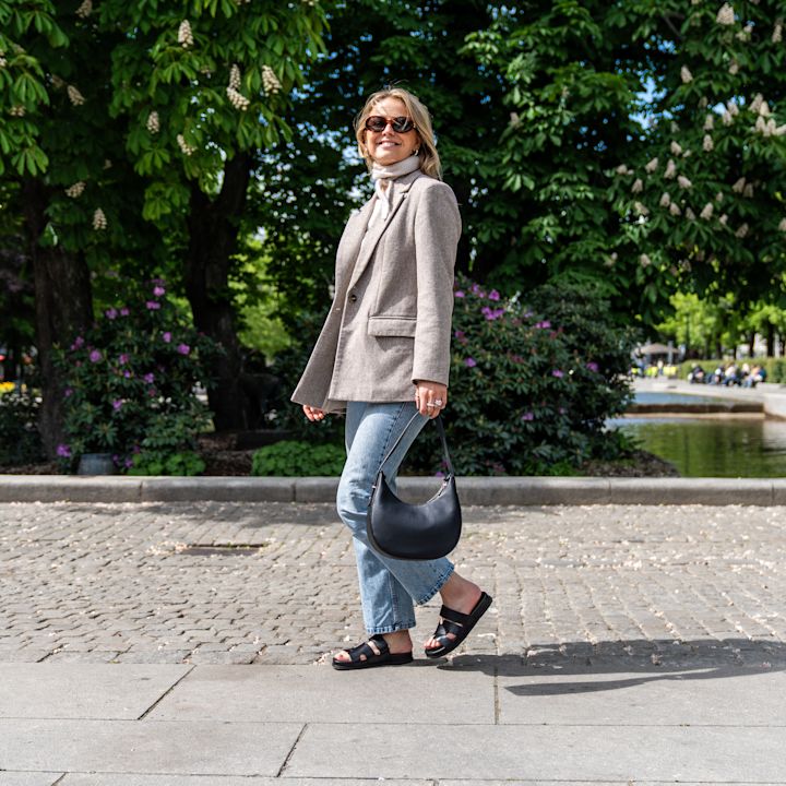 Person i beige jakke og jeans med svarte, åpne sandaler i lær og en svart veske, poserer i en frodig park.