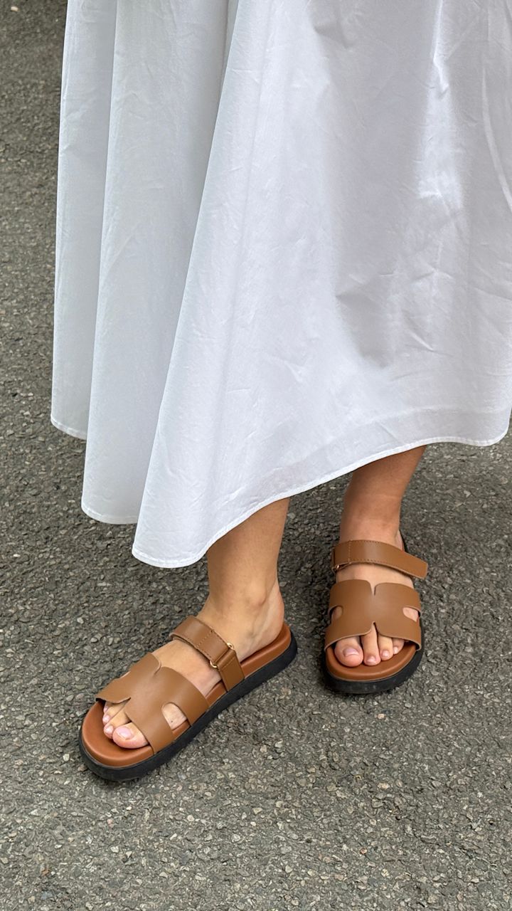Bruna sandaler med svart sula på fötter med vit lång klänning som står på asfalt.