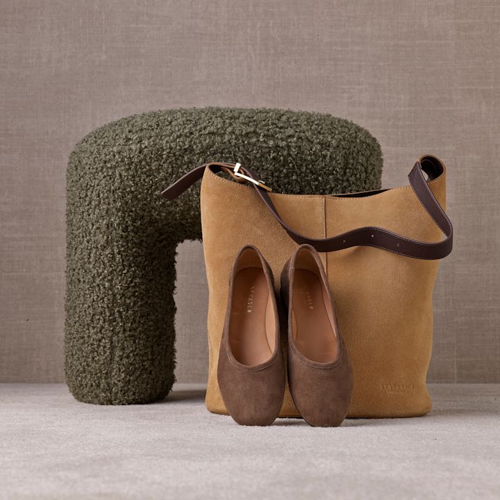 Bruna mockaballerinaskor framför en kamelbeige läderväska, placerade mot en grön textilpall med neutral bakgrund.