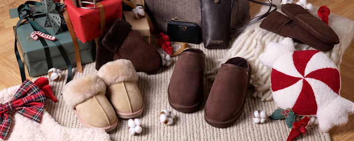 Mjuka tofflor i beige och brunt placerade på en stickad matta med julklappar, röd rutigt rosett och kuddar dekorerade med pepparmynta runt omkring.