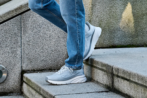 Person i blå jeans går uppför betongtrappa i ljusgrå sneakers från Okaki med vit sula.