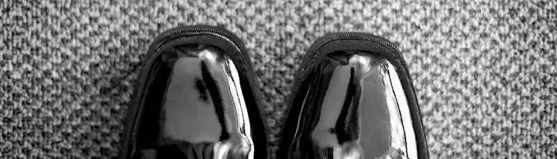 Svarta, blanka lackskor placerade på en grå, mönstrad matta sedd uppifrån.