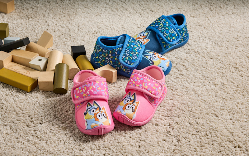 To par blå tøfler med tegneseriefigurer, en rosa og en blå, plassert på et lyst teppe ved siden av flere treklosser.