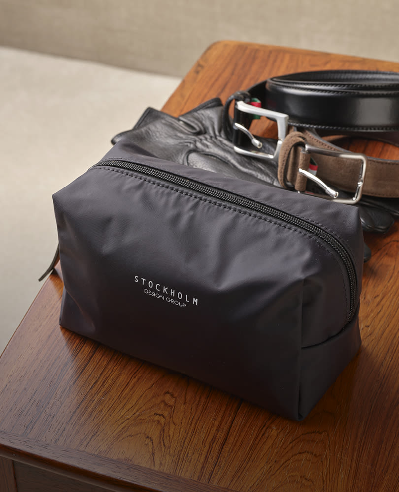 Svart toalettmappe fra Stockholm Design Group på trebord med svart og brun lærbelter ved siden av.