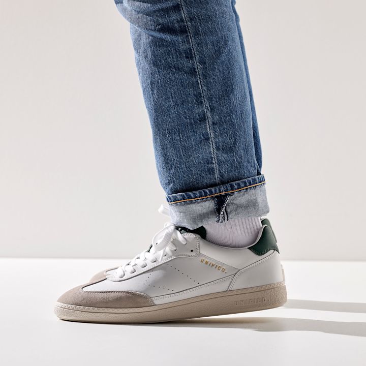 Hvit og beige Unified-sneaker med grønne detaljer, iført hvite sokker og blå jeans som er brettet opp nederst, står på hvit bakgrunn.