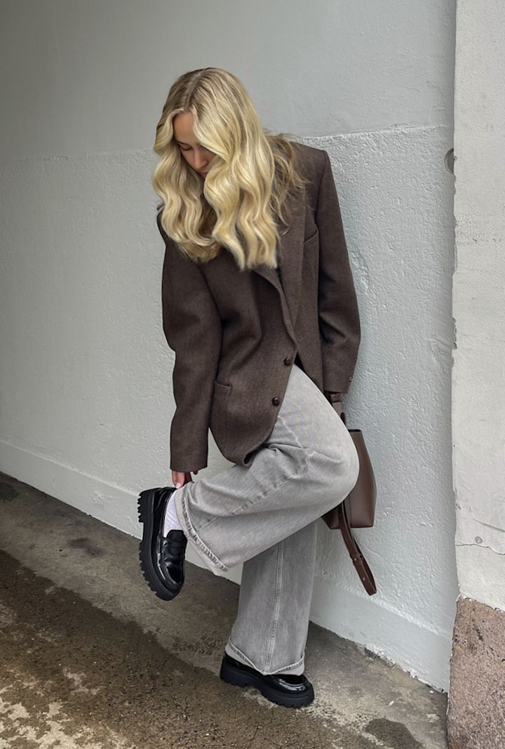 Kvinna med långt blont hår böjer sig ner och justerar sin sko medan hon står vid en vit vägg utomhus.