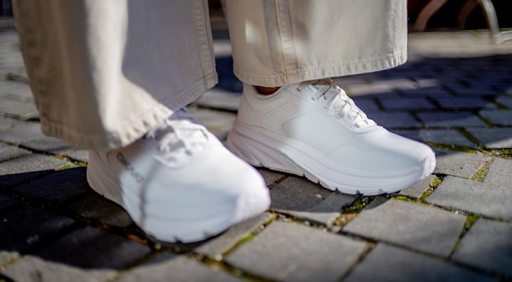 Hvite, moderne joggesko med tykk såle kombinert med lyse bukser, fotografert på brosteinsunderlag.