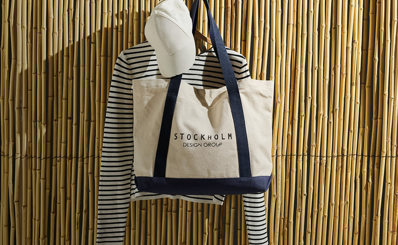 Hvit caps og stripet genser hengt med en beige og marineblå Stockholm Design Group-toteveske mot bambusvegg.